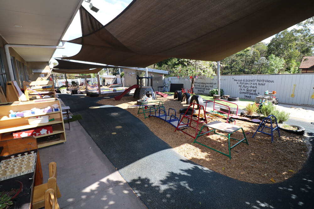 Large outdoor childcare playground with climbing equipment, natural surfaces and shade sails supporting active early learning.