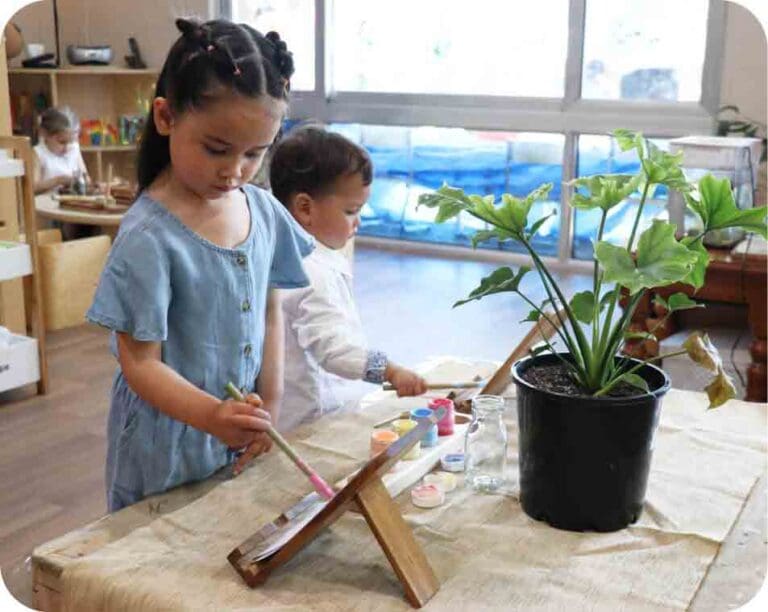 Children engaging in creative activities at Starfish Early Learning Centre, one painting on an easel and another observing, with a potted plant nearby.