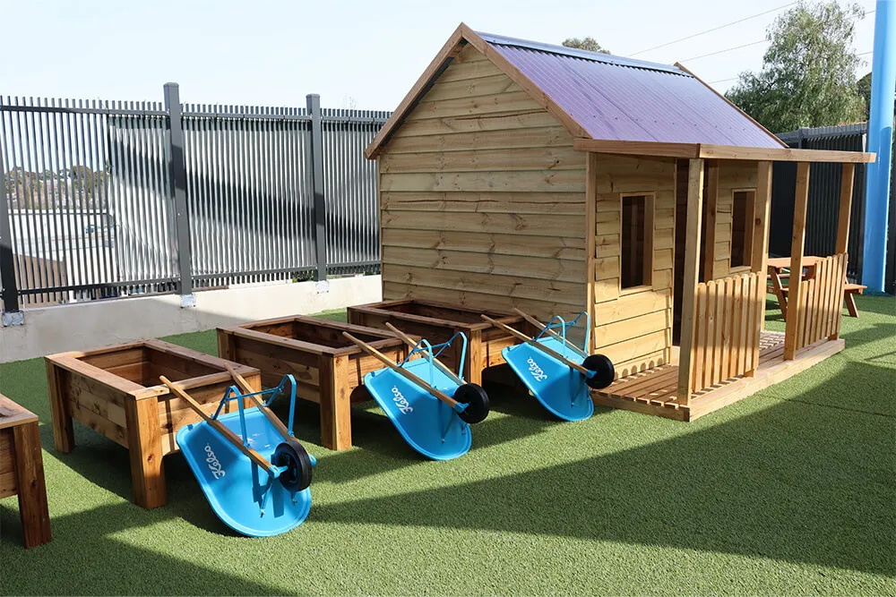 Outdoor childcare cubby house with garden beds and wheelbarrows supporting hands-on early learning play.