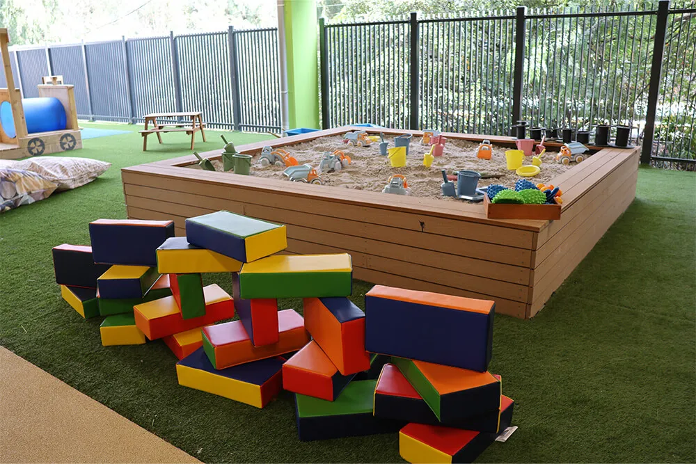 Outdoor childcare sandpit with toys and soft building blocks supporting hands-on early learning play.