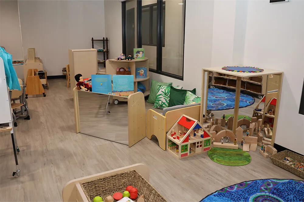 Childcare dramatic play and construction area with dolls, books and wooden blocks supporting early learning.