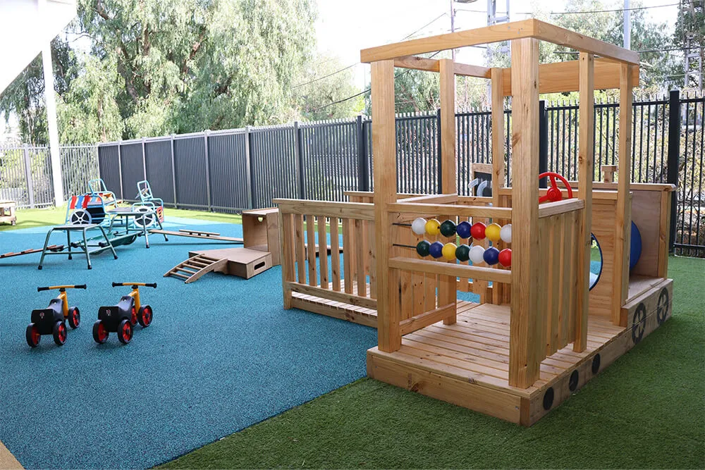 Outdoor childcare playground with wooden cubby structure, ride-on bikes and balance equipment for active early learning.