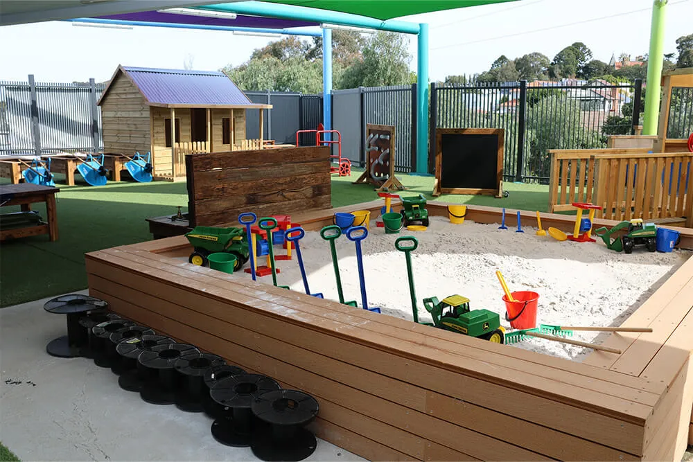 Outdoor childcare sandpit play area with shovels, trucks and cubby house for hands-on early learning.
