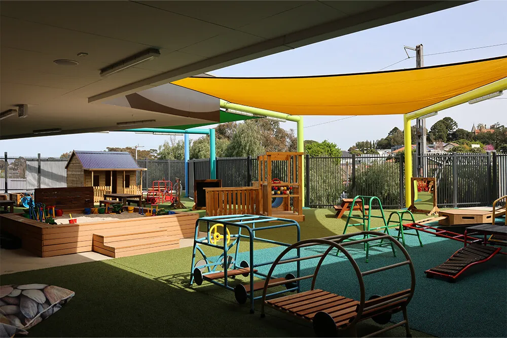 Large shaded outdoor childcare playground with climbing frames, balance equipment and play areas for active early learning.