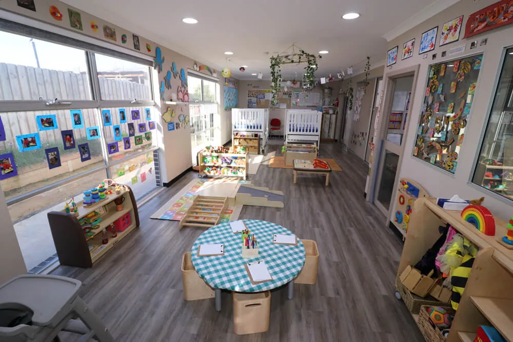 Bright childcare classroom with learning tables, artwork displays and play areas in a kindergarten early learning environment.