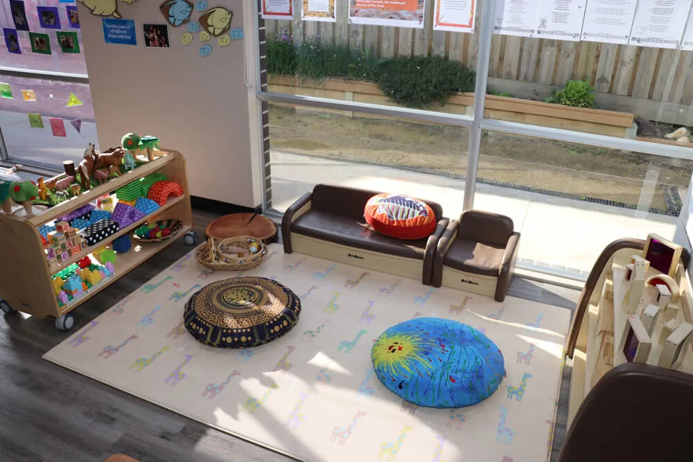 Cosy childcare reading and relaxation corner with cushions, soft seating and toys in a bright early learning classroom.