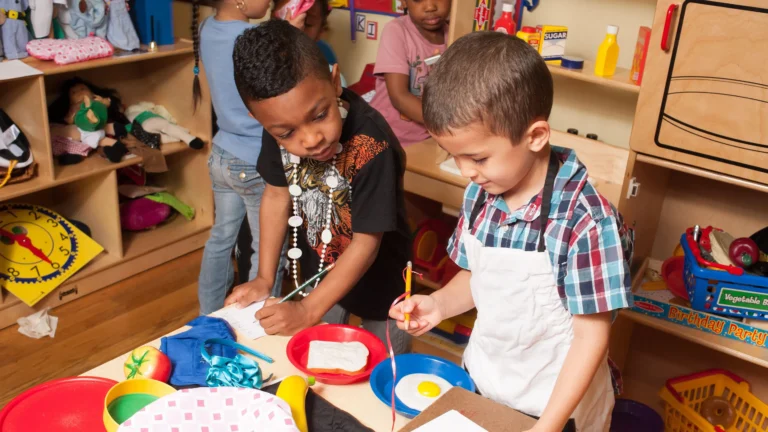 Children engaging in imaginative play together in an early learning classroom, exploring pretend food and writing materials at a play kitchen station.