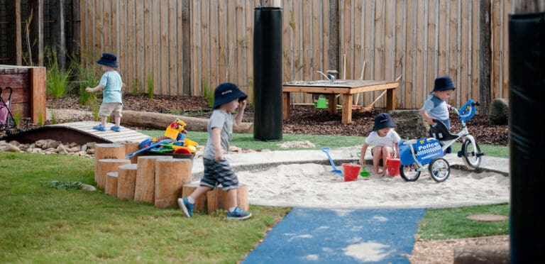 Children playing in a sandpit and riding toys in a childcare outdoor play area.