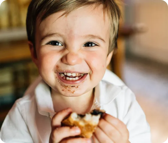 Smiling young child with messy chocolate around the mouth holding and eating a snack.
