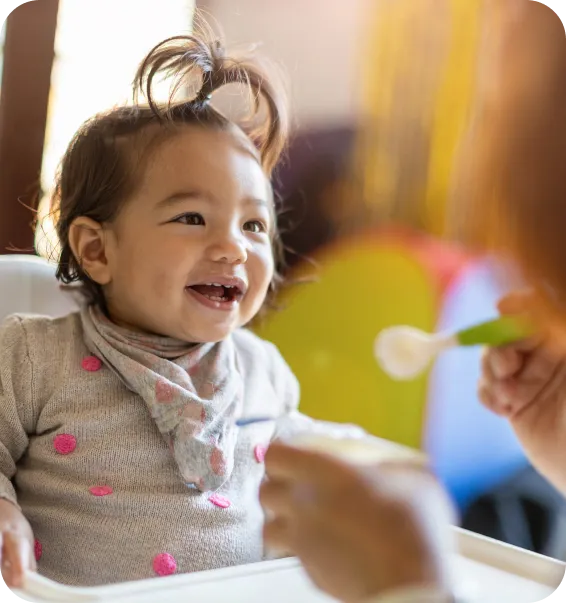 Smiling toddler being fed with a spoon during snack time in a childcare centre supporting safe early learning.