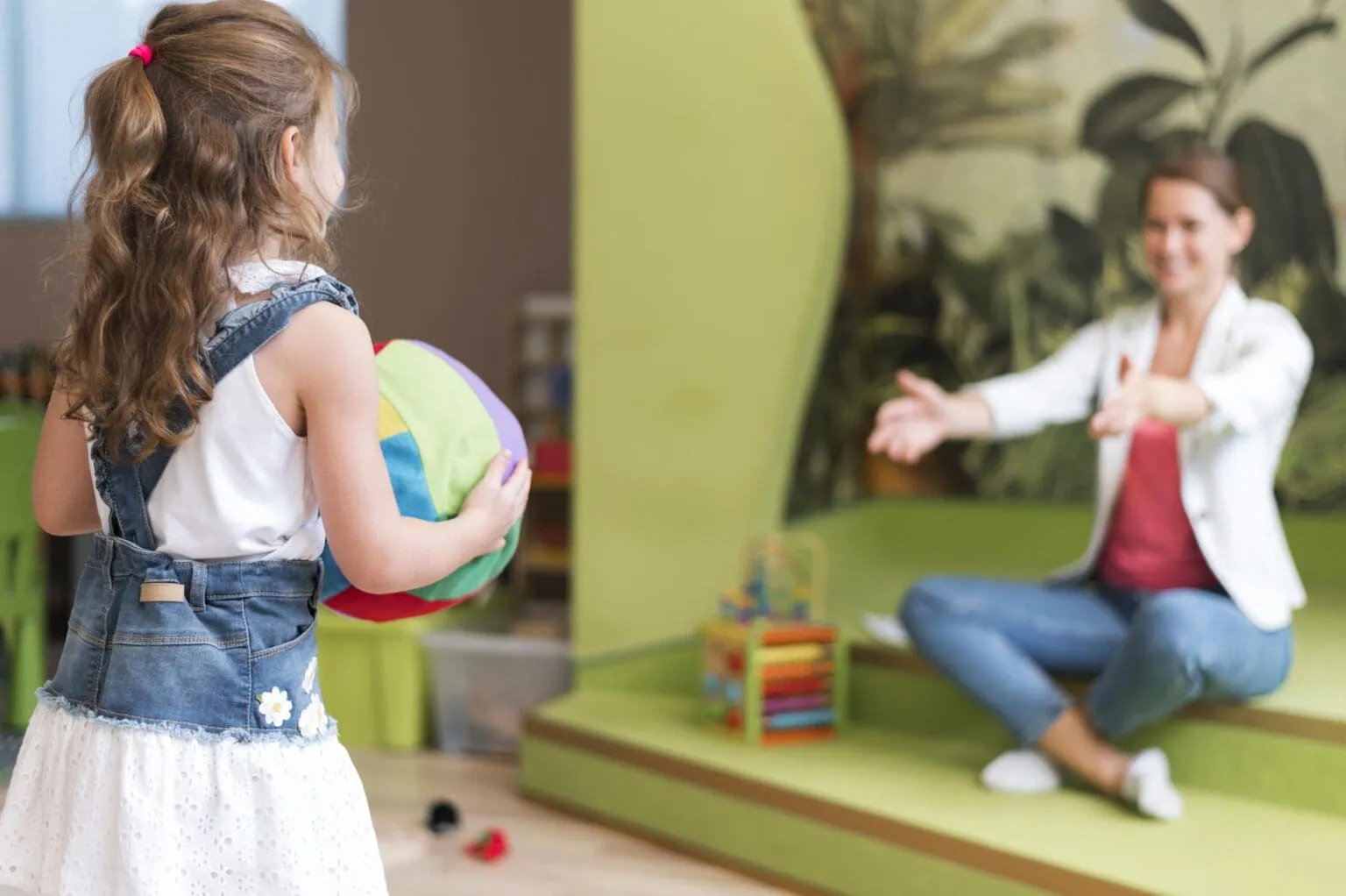 Child holding a colourful ball while an educator encourages play during a play based childcare activity.