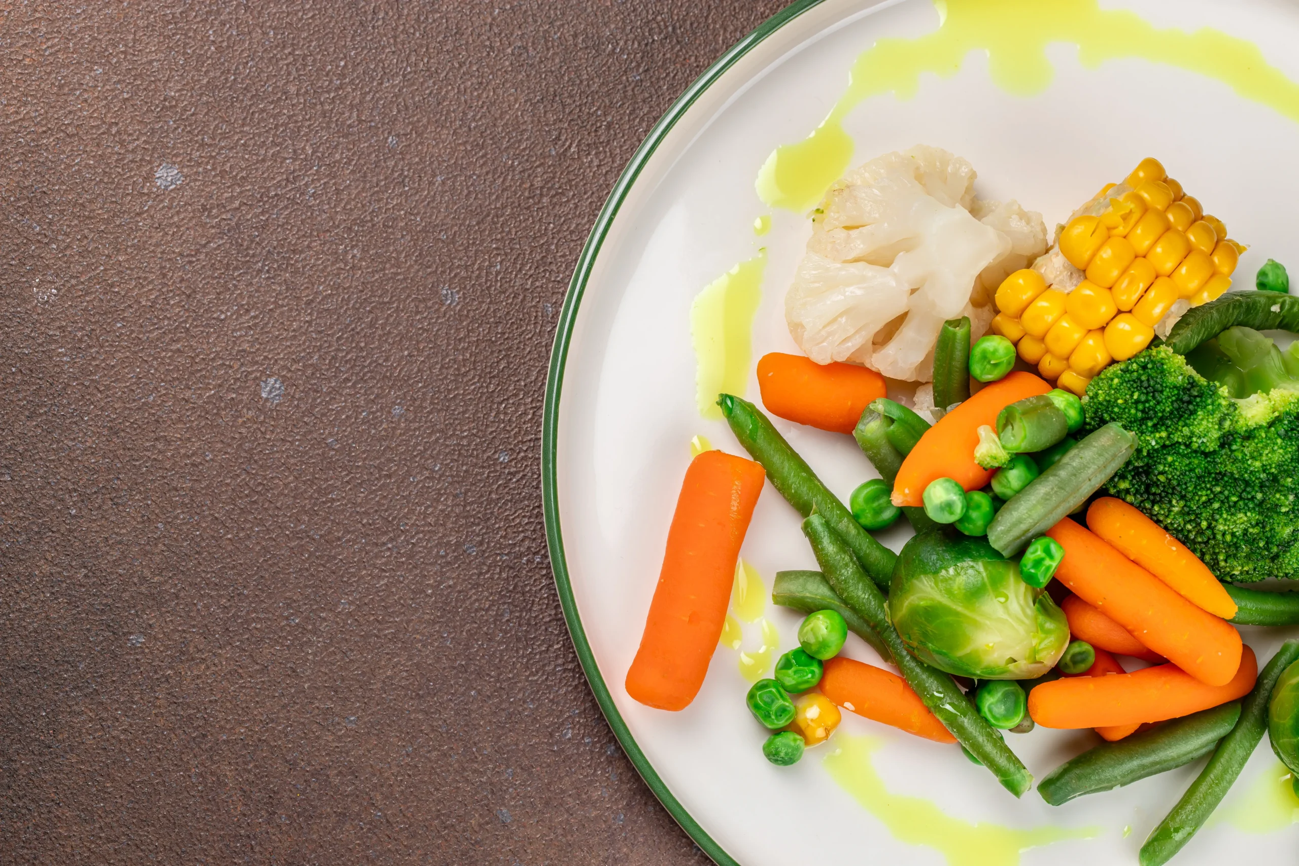Plate of colourful steamed vegetables, including carrots, broccoli, corn, peas and green beans.